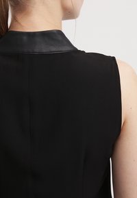 Svart ärmlös plagg med en kontrasterande krage i svart läder. Slät tygtextur, figurnära passform och rena designlinjer.