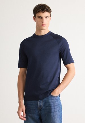 Mann mit kurzen, lockigen Haaren trägt ein dunkelblaues Kurzarmhemd und eine blaue Jeans. Er steht mit einer Hand in der Tasche vor einem schlichten Hintergrund.