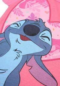 T-shirt en tissu rose avec un personnage de dessin animé bleu aux grands yeux et à l'expression souriante, entouré de motifs floraux.