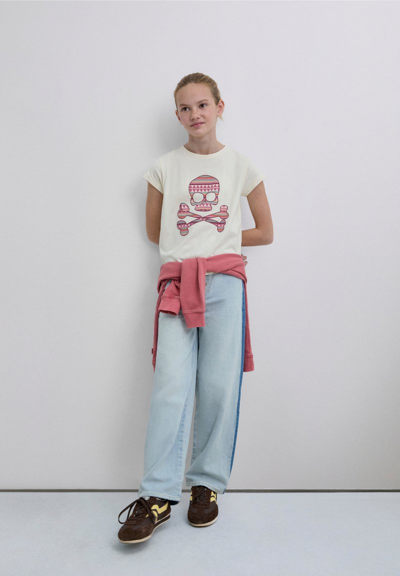 Weißes T-Shirt mit kurzen Ärmeln und einem bunten Totenkopf-Print, kombiniert mit hellblauen Jeans mit weitem Bein und braunen Sneakern mit gelben Akzenten. Rosa Sweatshirt um die Taille gebunden.