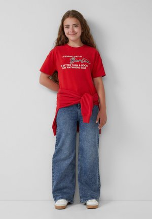Rotes Baumwoll-T-Shirt mit weißen Text, lässige Passform, kombiniert mit lockeren blauen Jeans. Sneakers mit hellen Sohlen und einem über das Hemd gebundenen Detail.
