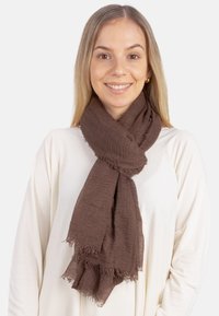 Écharpe marron texturée avec des bords effilochés, drapée autour du cou, portée sur un haut blanc. Matière douce et légère qui apporte de la chaleur.