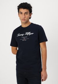 Granatowa bawełniana koszulka z krótkim rękawem, z białym napisem "Tommy Hilfiger" oraz rokiem powstania na przodzie.