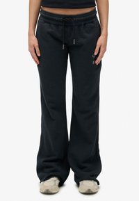 Persoon in zwarte wijde sweatpants met een trekkoord in de taille en beige sneakers, staande tegen een effen witte achtergrond.