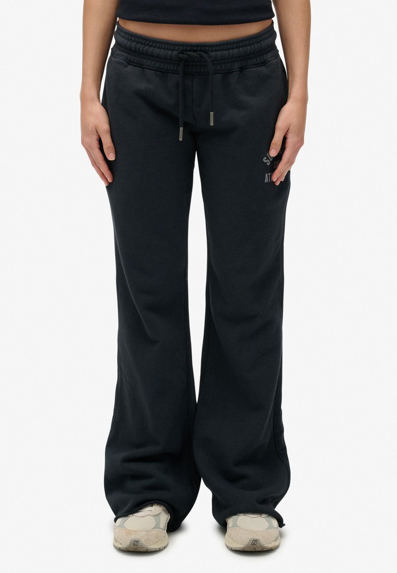 Persoon in zwarte wijde sweatpants met een trekkoord in de taille en beige sneakers, staande tegen een effen witte achtergrond.