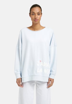 Woman facing forward wearing a loose, light blue sweatshirt with white text "J’adore la mer" and wide white pants, neutral background.