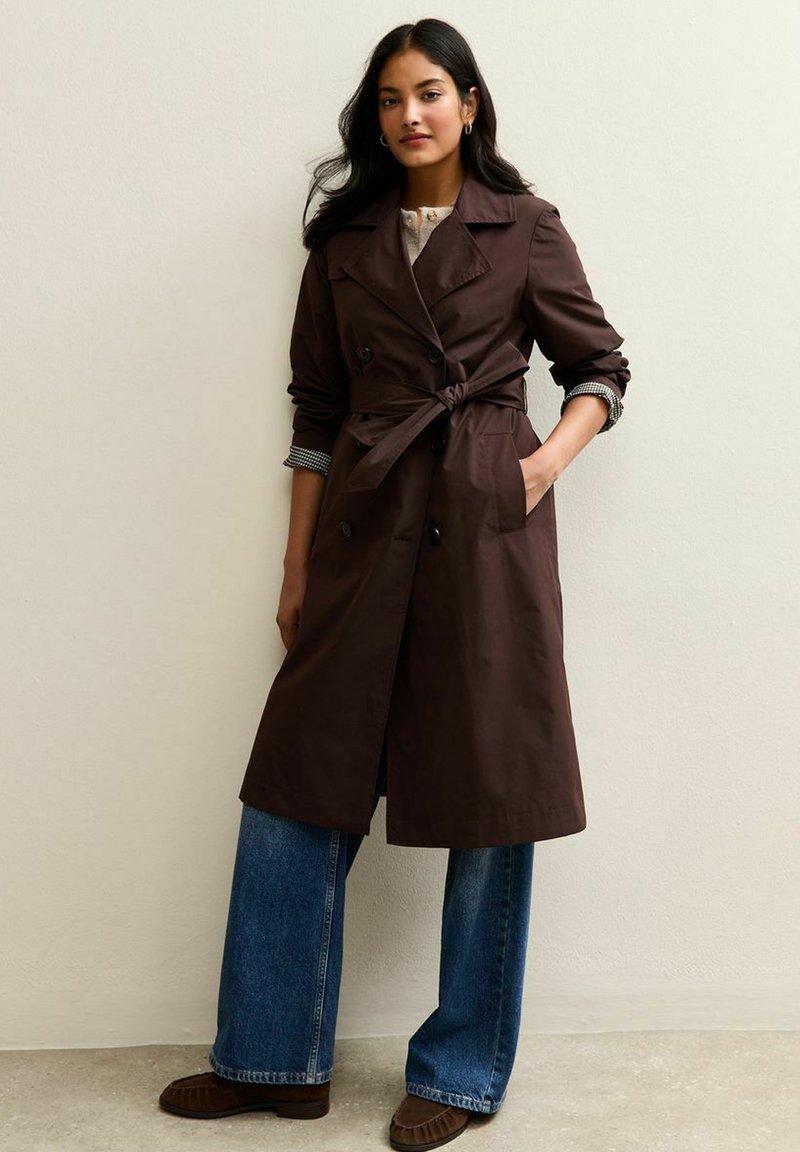Cappotto beige con cintura in vita, bottoni doppi, e maniche arrotolate su una camicia a quadri, abbinato a jeans a gamba larga.