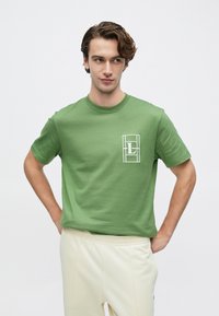 Πράσινο κοντομάνικο μπλουζάκι από απαλό βαμβάκι· διαθέτει λευκή επιγραφή με γράμματα σε ορθογώνιο πλαίσιο στο αριστερό στήθος.