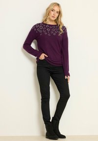 Kvinna i en lila tröja med strassleopardfläckar, svarta smala jeans och svarta ankelstövlar, stående med ena handen i fickan.