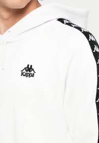 Sudadera blanca con cordón y logotipo negro de Kappa en el pecho. Estrías laterales negras con logotipos de Kappa en blanco que se repiten. Textura suave.