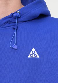 Μπλε φούτερ με ανάγλυφο ύφασμα. Διαθέτει κλείσιμο με κορδόνια, ασημένια εξαρτήματα και λευκό τριγωνικό λογότυπο με σχέδιο "a CG".