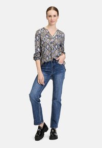 Hemd mit Knopfleiste und geometrischem Muster in Blau, Gelb und Braun; kombiniert mit blauen Jeans; schwarze Plateau-Schuhe; Hände in den Taschen.