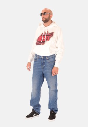 Witte hoodie met rode afbeelding, blauwe jeans en zwarte sneakers. Ronde zonnebril en een neutrale achtergrond maken de outfit compleet.