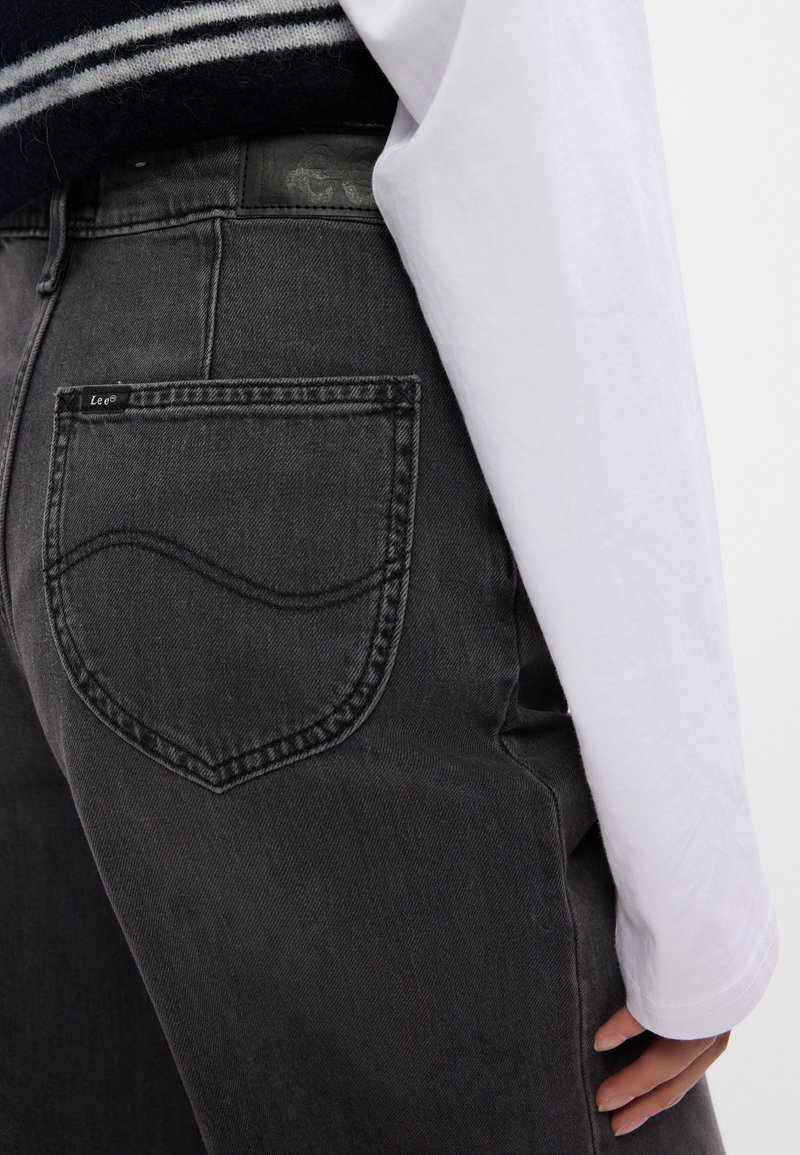 Zbliżenie na osobę w czarnych dżinsach Lee i białej koszuli z długim rękawem, ukazujące tylną kieszeń i część dłoni.