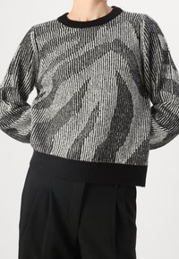 Randig svartvit tröja med en texturerad mönster, rundad svart halsringning och ribbat avslut, bärs över svarta byxor.