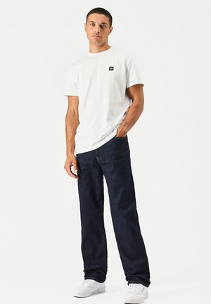 Junger Mann steht mit einer Hand in der Tasche, trägt ein weißes T-Shirt, dunkelblaue Jeans und weiße Turnschuhe vor einem schlichten Hintergrund.