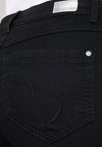 Vaqueros de denim negro con un bolsillo trasero que presenta un sutil diseño bordado y un acento de logo metálico cerca de la costura.
