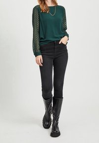 Dunkelgrünes Oberteil mit gemusterten transparenten Ärmeln, kombiniert mit schwarzen engen Jeans und kniehohen schwarzen Stiefeln, was einen Layered-Look erzeugt.