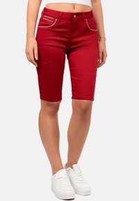Bermuda shorts in denim rosso con design aderente, caratterizzati da tasche frontali con accenti argentati e una texture liscia. Abbinati a sneakers bianche.