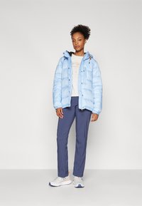 Giacca a molletta azzurra chiara con cappuccio, zip frontale e polsini elastici. Abbinata a pantaloni blu scuro e sneaker bianche con dettagli arancioni.