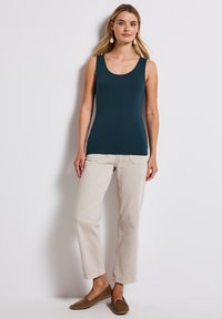 Top in cotone navy a design aderente abbinato a pantaloni beige chiaro dritti e scarpe senza lacci marroni, indossato su uno sfondo bianco semplice.