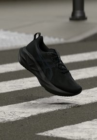 Μαύρο αθλητικό παπούτσι με υφασμένο επάνω μέρος, κομψό σχέδιο και ελαφρώς αντανακλαστικό λογότυπο, τοποθετημένο μπροστά σε φόντο διάβασης πεζών.