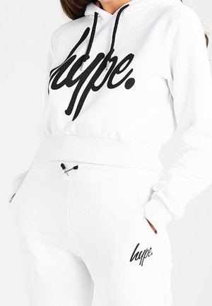 Nainen pukeutunut valkoiseen lyhyeen huppariin ja vastaaviin valkoisiin verkkareihin, joissa musta "hype." logo rinnassa ja lahkeessa.