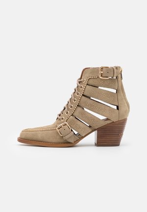 Taupe ruskind ankle boot med udskårne remme, snørelukning foran, gyldne nitter, to spænder og en stablet træblokhæl.