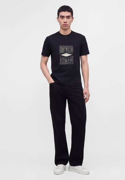 Νέος άνδρας που φοράει μαύρο T-shirt Emporio Armani, μαύρο παντελόνι και άσπρα αθλητικά παπούτσια, στέκεται με το ένα χέρι στην τσέπη σε λευκό φόντο.