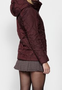 Doudoune bordeaux avec capuche, présentant un design matelassé et des poches latérales. Portée avec une jupe plissée à motif.