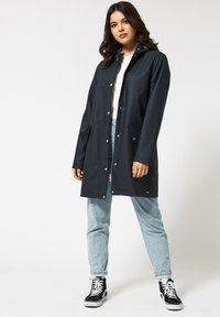 Tummansininen vedenpitävä sadetakki, jossa on etupainikkeet, huppu ja kaksi sivutaskua, päällä vaaleansiniset farkut ja mustat korkeakärkiset tennarit.