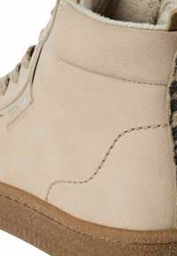 Bottine en daim beige avec des détails de coutures, un col rembourré et une semelle en caoutchouc brun texturé.