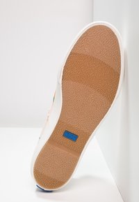 Semelle d'une basket blanche avec une semelle en caoutchouc brun clair texturée et un petit logo rectangulaire bleu Keds près du talon.