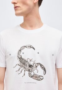 Άνδρας που φοράει λευκό μπλουζάκι με λεπτομερή μαύρη εικονογράφηση σκορπιού και διάσπαρτα γράμματα "H U G O" μπροστά.