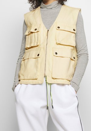 Person, die eine hellbeige Arbeitsweste mit Vorderfächern und Reißverschluss über einem grauen Langarmshirt und weißen Hosen mit schwarzen Seitenstreifen trägt.