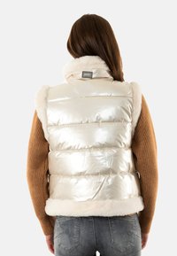 Gilet a palloncino iridescente con colletto e orlo in pelliccia, caratterizzato da trapuntatura orizzontale e un sottile logo sul retro. Completato da un maglione marrone.