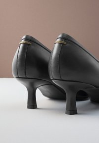Chaussures à talons hauts en cuir noir avec un bout arrondi et un talon carré bas. Accents dorés à l'arrière, texture lisse et design minimaliste.