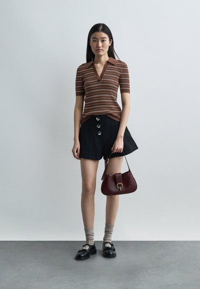 Jeune femme portant un haut à manches courtes rayé marron, un short noir à boutons sur le devant, des chaussettes beiges, des mocassins noirs, tenant un sac à main bordeaux.