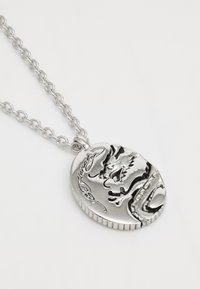 Srebrna zmajčina obesek na verigi, z obsežnim graviranjem in teksturirano površino. Ovalna oblika z zapletenim dizajnom.