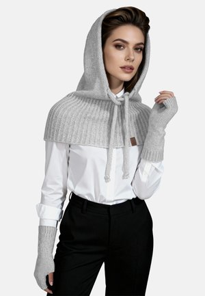 Grijze gebreide capuchoncape met geribbelde textuur en trekkoordsluiting, gecombineerd met vingerloze handschoenen. Draagt over een witte shirt en zwarte broek.