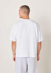 Bela bombažna majica s sproščenim krojem, okroglim izrezom in kratkimi rokavi, kombinirana s sivimi trenirkami. Preprost in minimalističen dizajn.