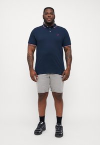 Námornícky polo tričko s červeným a bielym lemom, sivé šortky, čierne tenisky a ponožky po členky; model stojí na bielom pozadí.