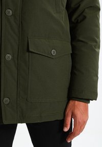 Olivgrüne Jacke mit durchgehendem Knopfverschluss, strukturiertem Stoff, großer Klappentasche und verstellbaren Manschetten; verfügt über fünf Knöpfe am Verschluss.
