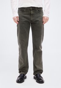 Άτομο που φοράει σκούρα ελιά τζιν με ευθεία γραμμή, λευκό πουκάμισο με μακριά μανίκια και μαύρα δερμάτινα μοκασίνια, στέκεται μπροστά σε ένα απλό ανοιχτό φόντο.