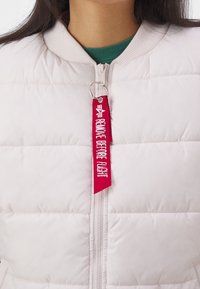 Helle rosa Pufferjacke mit einem gerippten Kragen, Frontreißverschluss und einem roten Stoffetikett mit der Aufschrift "VOR FLUG ENTFERNEN." Verfügt über gesteppte Abschnitte.