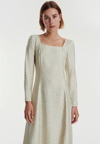 Hellgrünes Kleid mit quadratischem Ausschnitt, langen Ärmeln und einem dezenten, weißen Sprenkelmuster. Glattes Material mit leichtem Glanz.