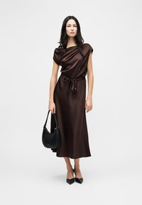 Γυναίκα με καφέ μεταξωτό midi φόρεμα με ζώνη στη μέση, κρατώντας μαύρη τσάντα χειρός και φορόντας μαύρες μυτερές γόβες, στέκεται μπροστά σε μια μονόχρωμη φόντο.