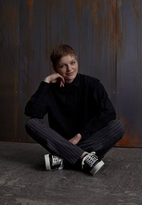 Persona seduta con le gambe incrociate su un pavimento texture, che indossa una camicia nera abbottonata e pantaloni grigi a righe, con sneakers bianche e nere.