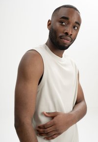 Ärmelloses, hellbeiges Oberteil aus weichem Stoff, mit rundem Ausschnitt und lockerer Passform. Das Model zeigt einen neutralen Gesichtsausdruck.