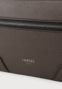 Καφέ τσάντα από δέρμα με υφή, με μεταλλικό φερμουάρ και εντυπωμένο λογότυπο της μάρκας "LANCEL PARIS" σε χρυσό στο κάτω σημείο.